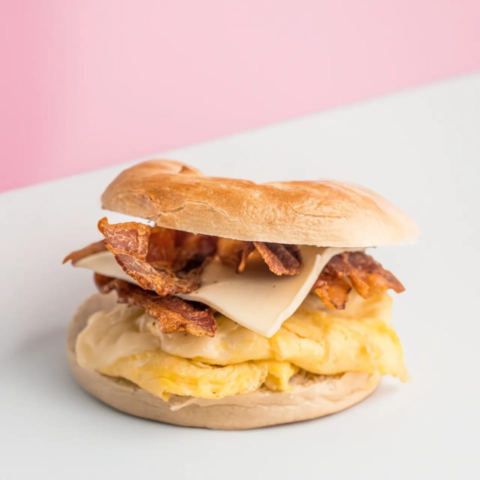 A breakfast sandwich with scrambled eggs, cheese, and bacon on a bagel against a pink and white background.