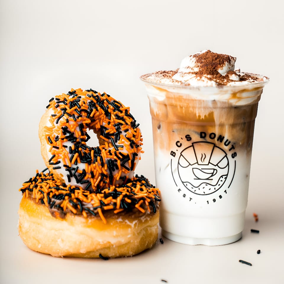 Two Halloween-themed donuts with orange and black sprinkles next to an iced coffee from B.C's Donuts.